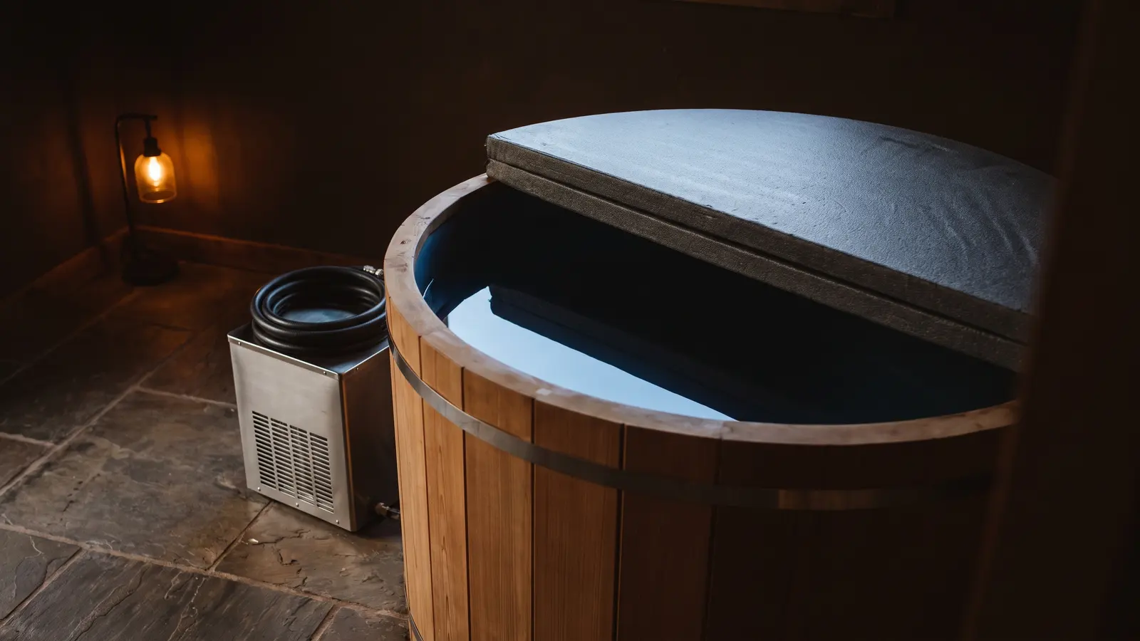 A cedar cold plunge tub with its insulated lid slid half off over still dark water, a stainless chiller unit with a coiled hose on the slate floor beside it, one small lamp on the far wall.