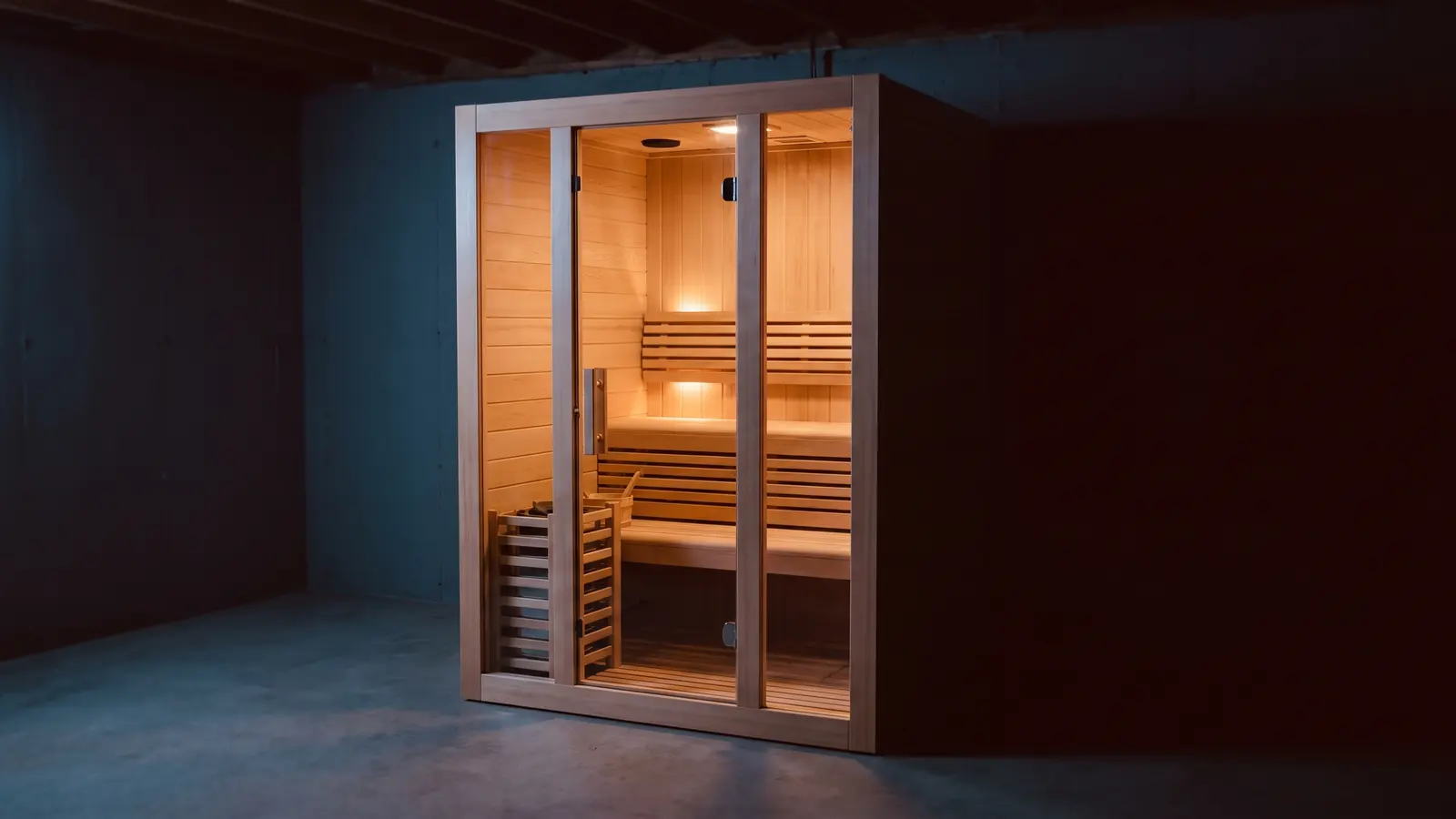 A freestanding infrared sauna cabin standing on a bare concrete floor in an unlit basement, its full glass front the only lit thing in the room, pale hemlock benches visible inside.